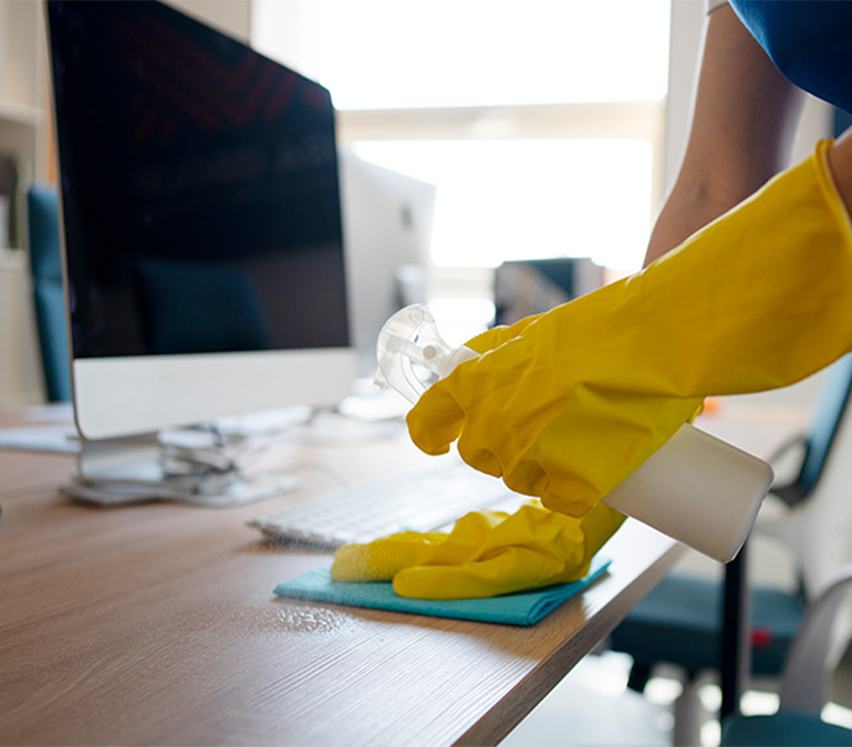 Close-up of a keeply employee sanitizing a wood-grain office desk next to a computer monitor.