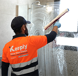 A Keeply employee cleaning a glass shower partition in a modern bathroom.