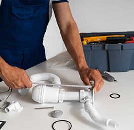 a keeply employee using a measuring tape to size a white plastic sink trap on a white table, with a grey toolbox in the background.
