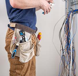 A keeply employee wearing a tool-filled belt and using pliers to work on a complex cluster of electric wires emerging from a wall electric box.